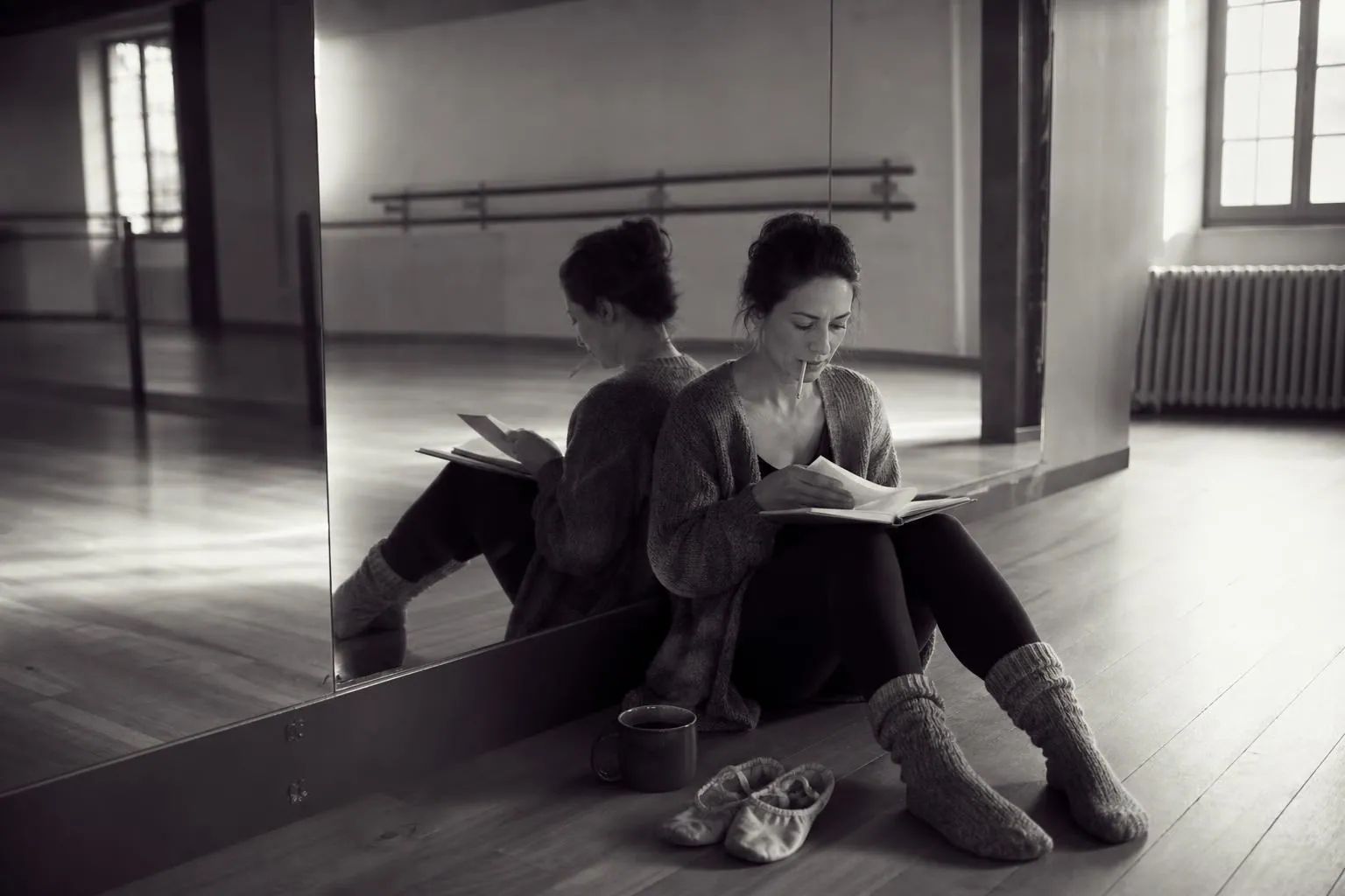 Une professeure de danse assise au sol contre le miroir d’un studio vide, un agenda papier ouvert sur les genoux, au petit matin.