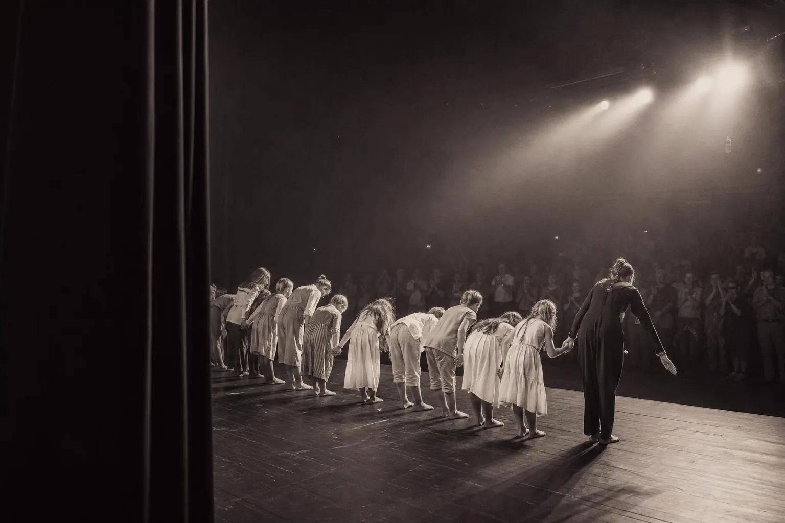 Vue de coulisse sur une rangée d’enfants en costume alignés face au public au moment du salut final, rideau ouvert et salle debout.