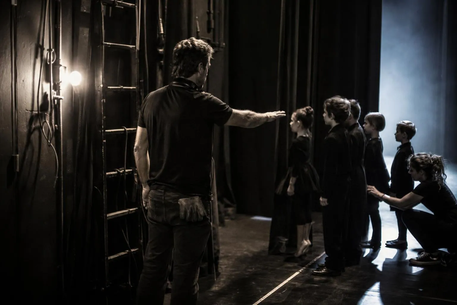 Un régisseur casqué d’intercom autour du cou retient un groupe d’enfants en costume derrière la ligne de sécurité en coulisse pendant un spectacle.