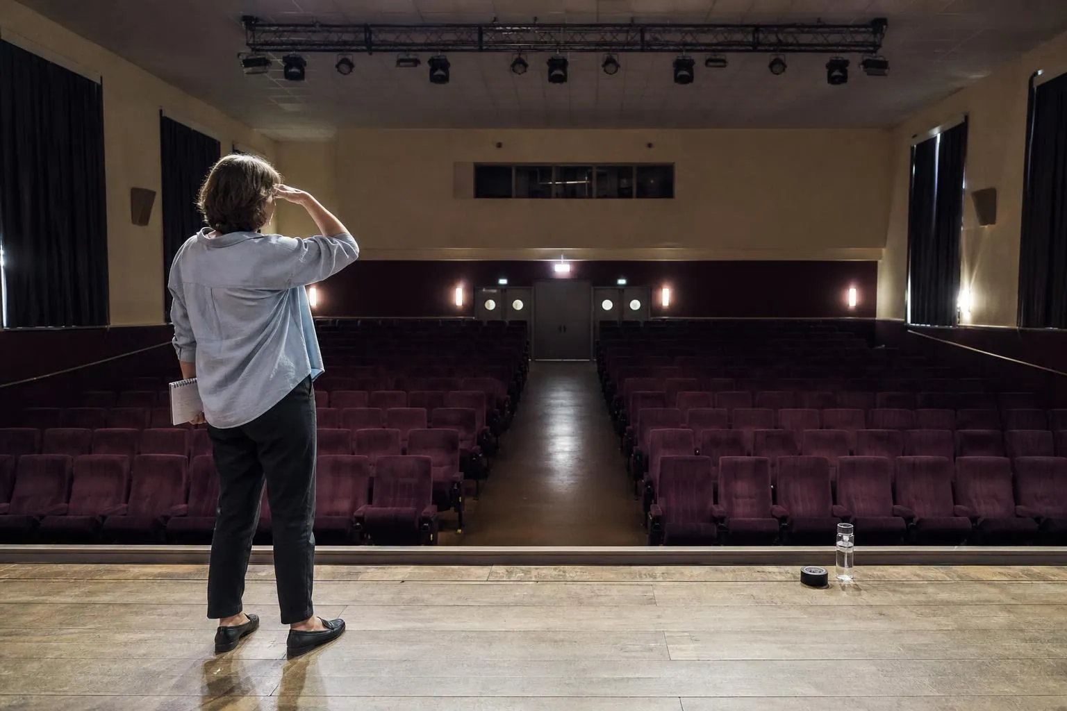 Une directrice d’école de danse debout au bord du plateau d’une salle des fêtes vide, regardant les rangées de fauteuils avant la mise en place du gala.