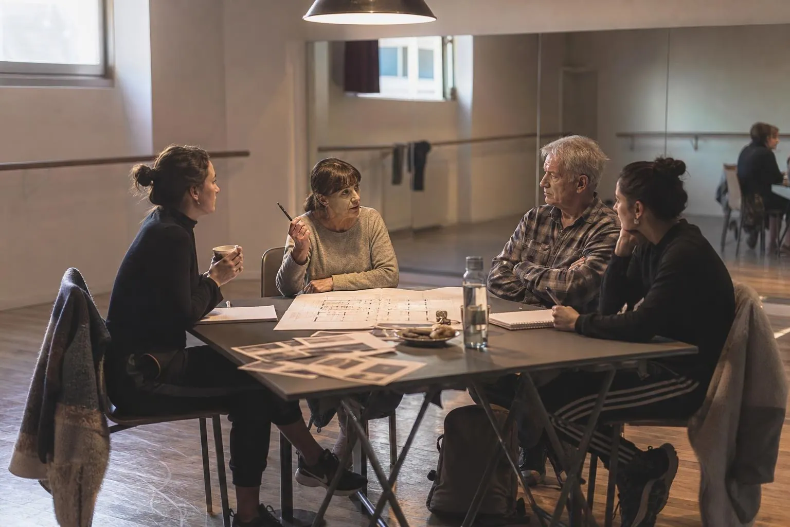 Quatre membres du bureau d’une école de danse réunis autour d’une table de studio couverte de plans de salle et de feuilles de préparation du gala.