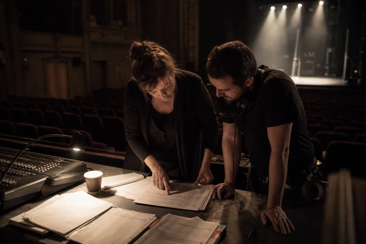 Une directrice d’école et un régisseur penchés sur une table de régie au fond d’une salle de spectacle pendant le filage, feuilles de conduite étalées